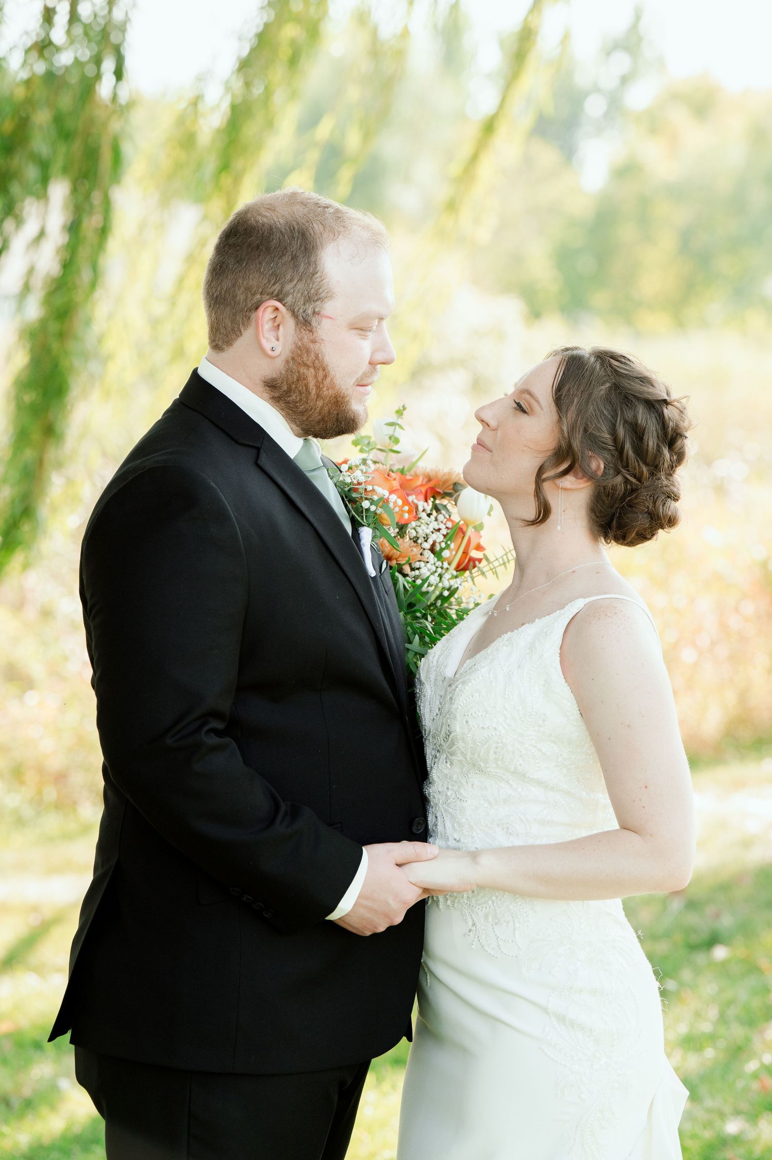 The Grands At Mulligans. A newlywed couple stands against a dreamy outdoor backdrop with willow trees and autumn foliage.