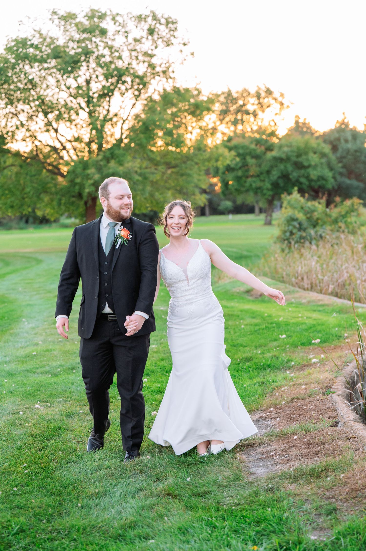 The Grands at Mulligans- Wedding Photos: Newlyweds stroll together on a grassy path during sunset.