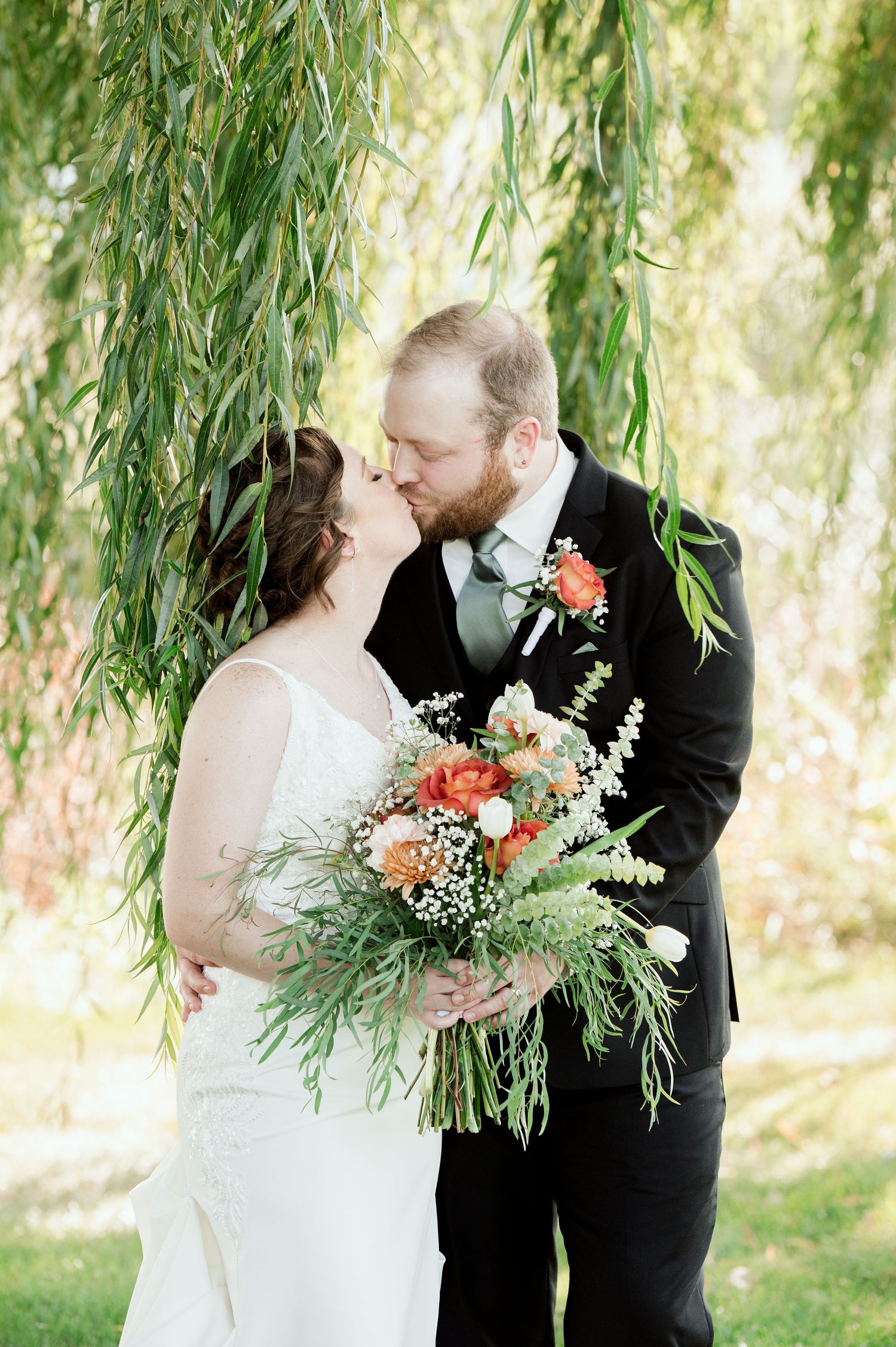 A romantic wedding couple shares a kiss under drooping willow branches with a lush floral bouquet.
