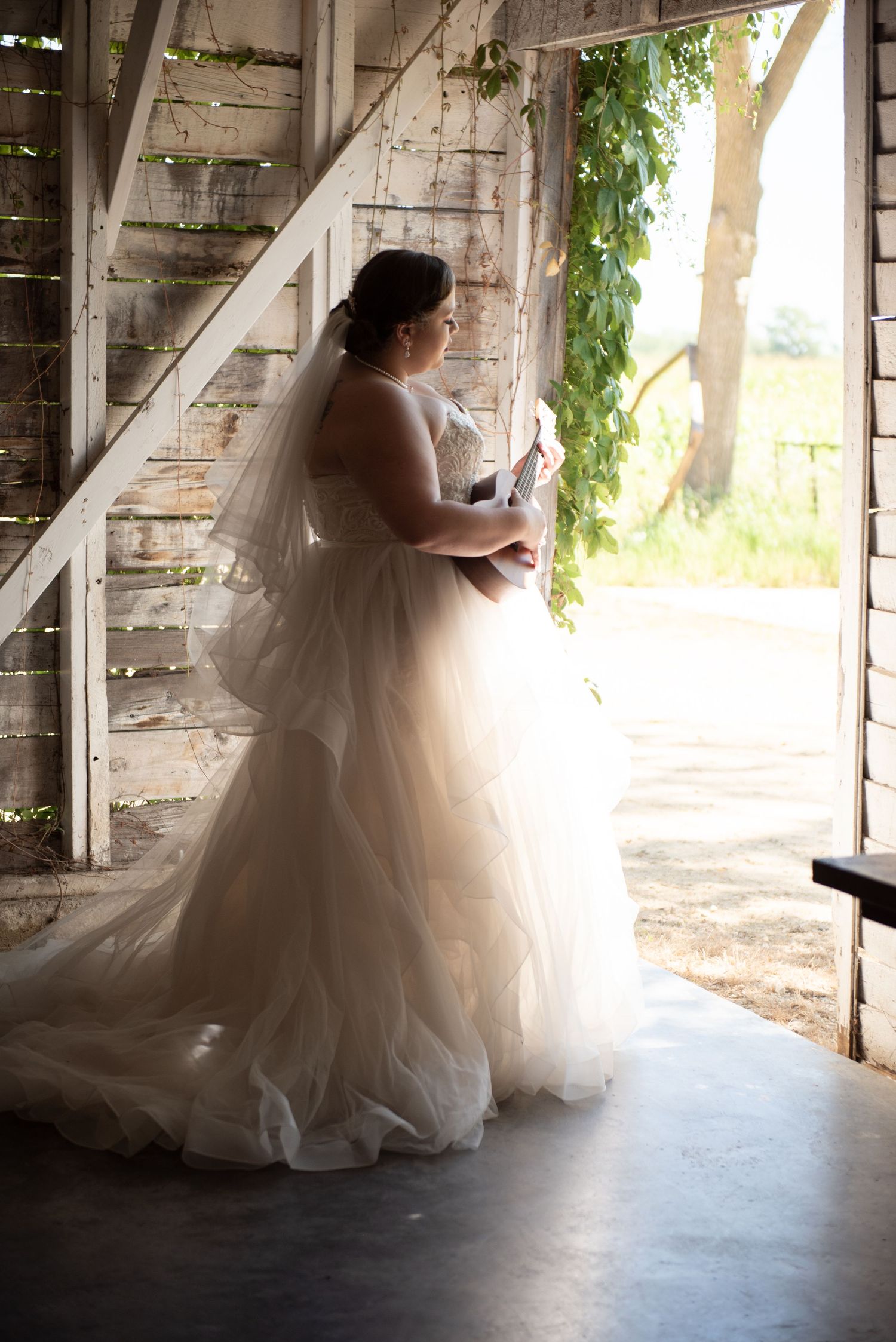 A bride in a flowing white tulle gown stands in silhouette by a rustic barn doorway with vines growing nearby.