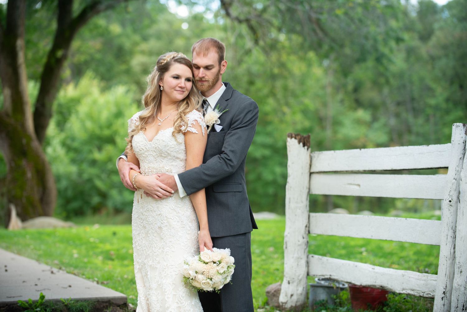 Romantic wedding portrait of a couple near a white fence with lush greenery in the background. Rum River Barn Milaca Minnesota Weddings