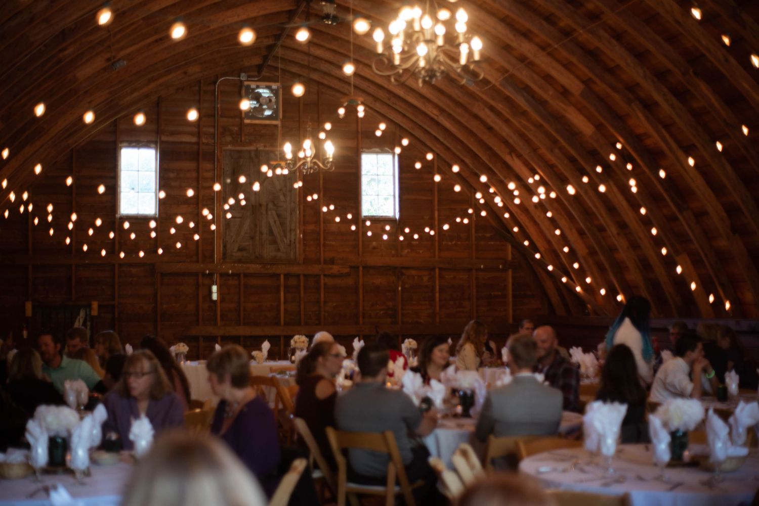 Reception guests dine beneath twinkling string lights and chandeliers inside a rustic wooden barn venue.