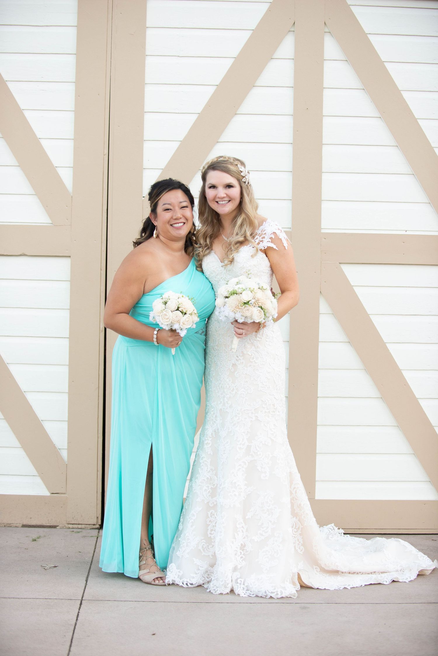 A bride in a white lace gown and bridesmaid in turquoise dress hold bouquets while posing against a white barn door. Milaca Minnesota Weddin