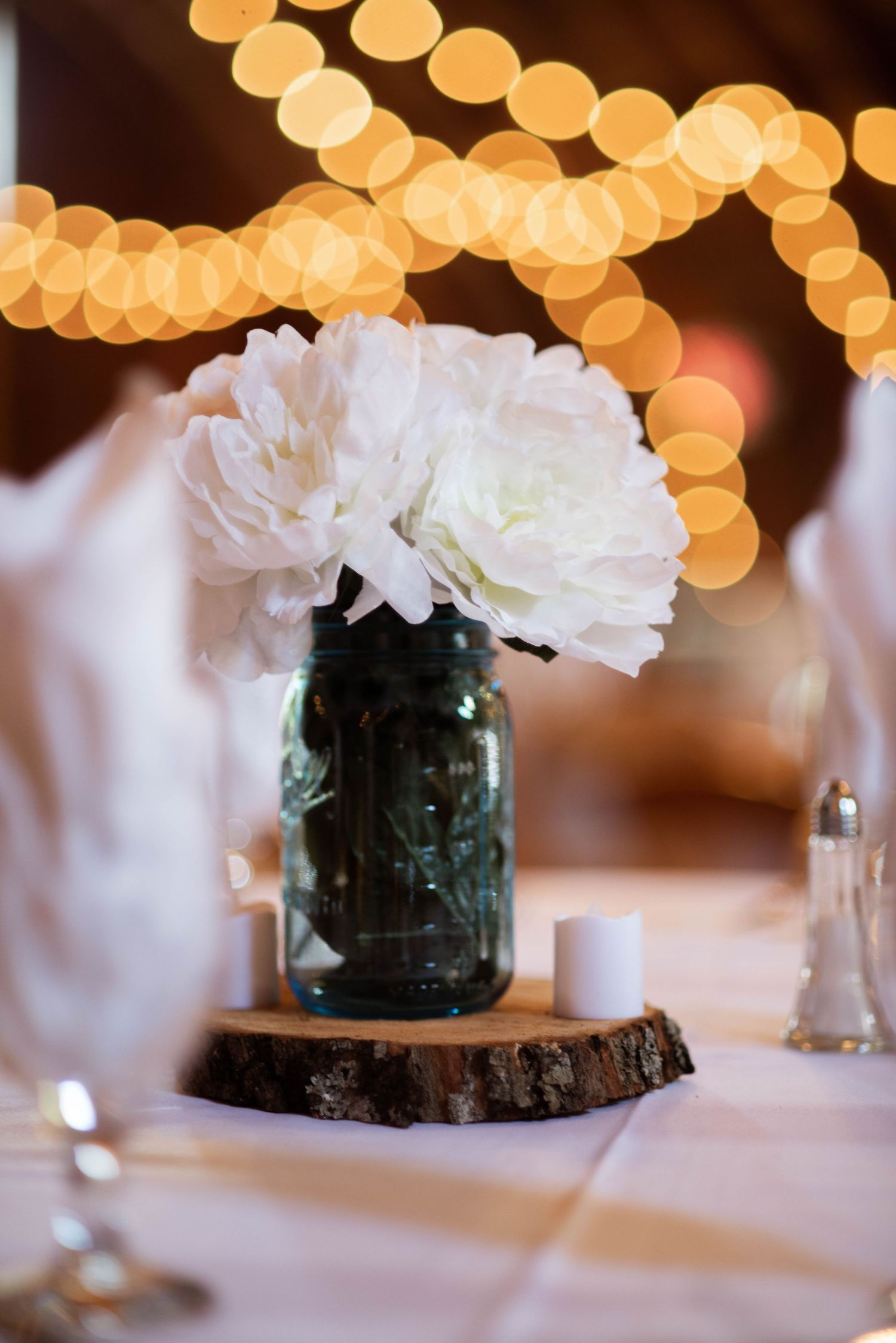 White carnations in blue mason jar centerpiece with bokeh lights creating warm ambiance in background.