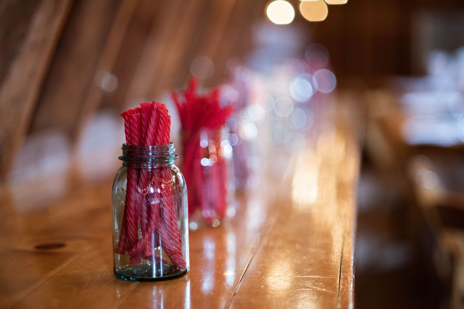 Red drinking straws in glass jars arranged on a wooden bar counter with soft bokeh lights in the background.