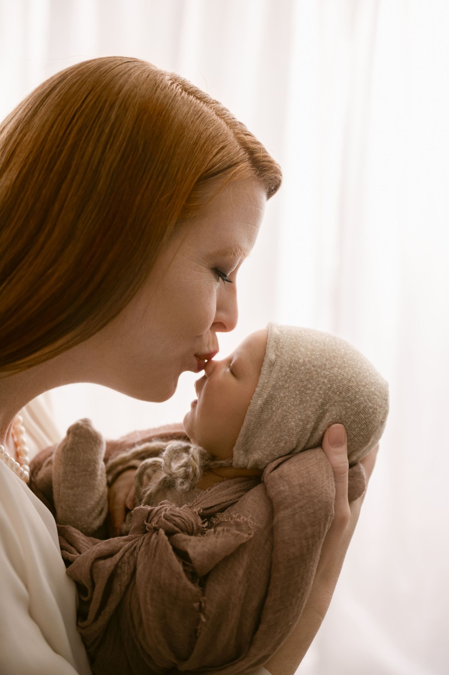 A mother in white gives her swaddled newborn baby a gentle kiss in a tender moment by a window.