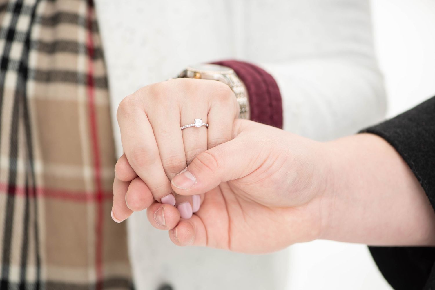 A sparkling diamond engagement ring is displayed on someone's hand against a plaid and white background.