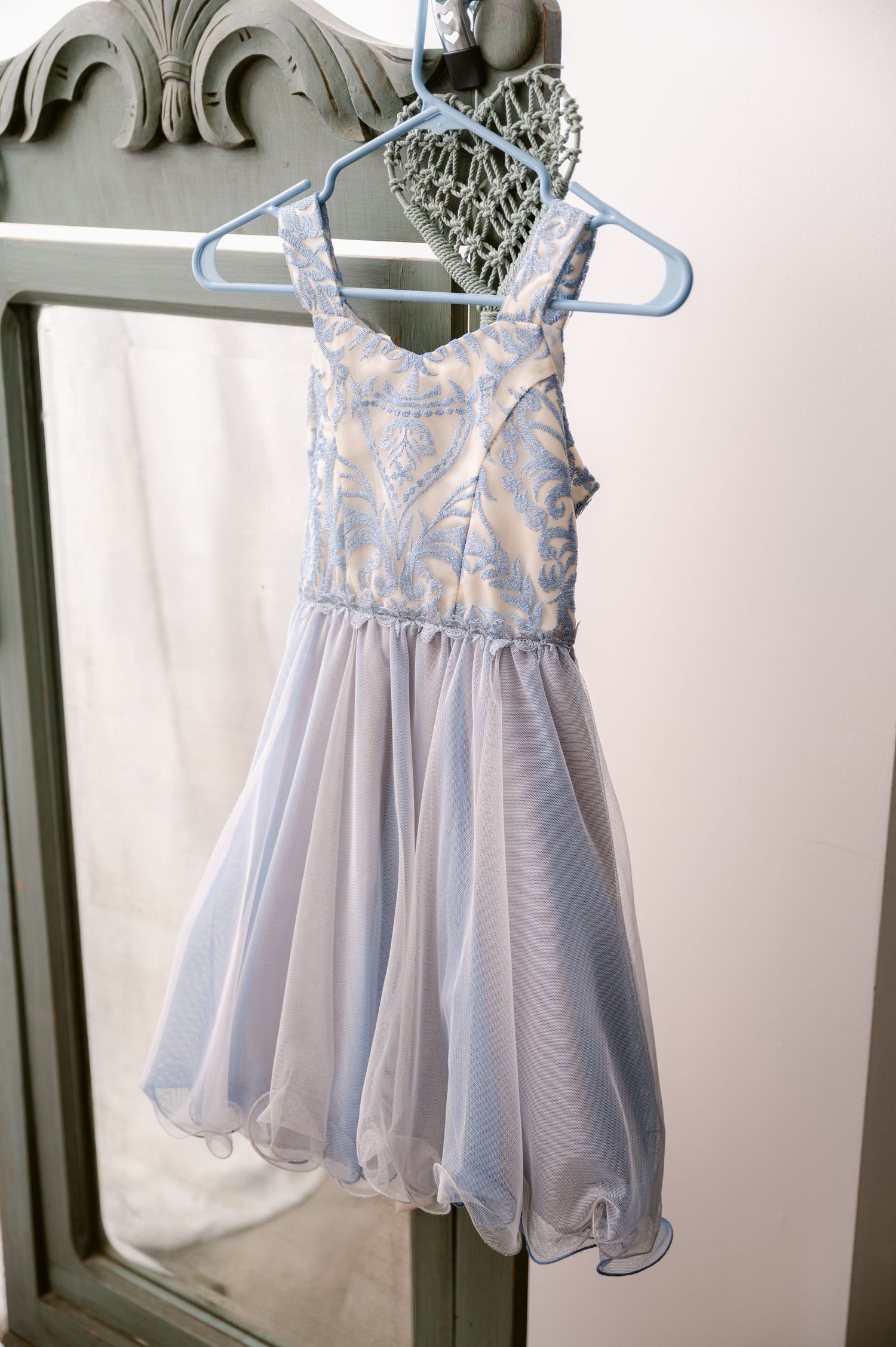 Light blue chiffon party dress with floral lace bodice and full skirt hanging on white plastic hanger against ornate mirror.