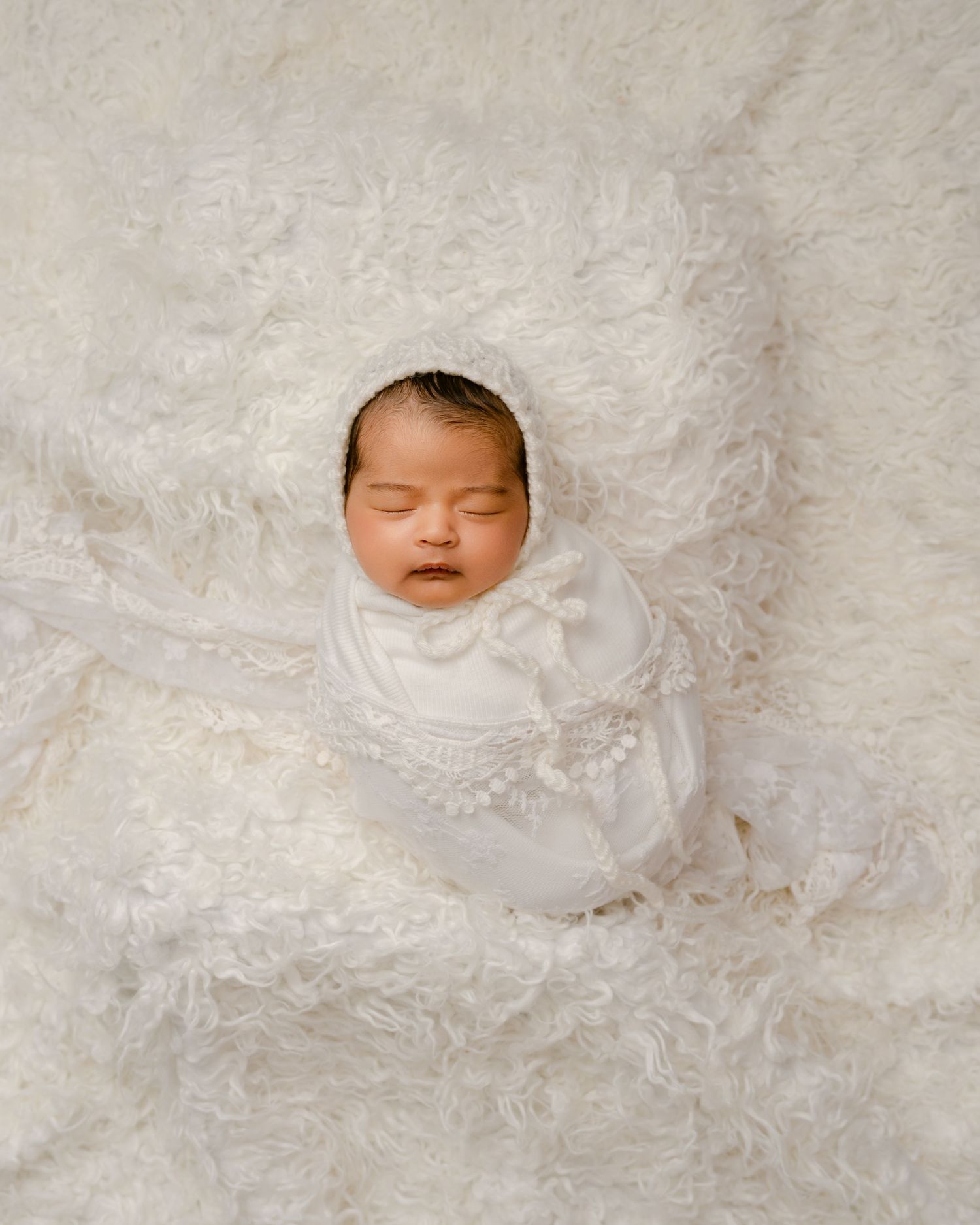 Peaceful sleeping newborn wrapped in white lace swaddle against creamy textured backdrop.