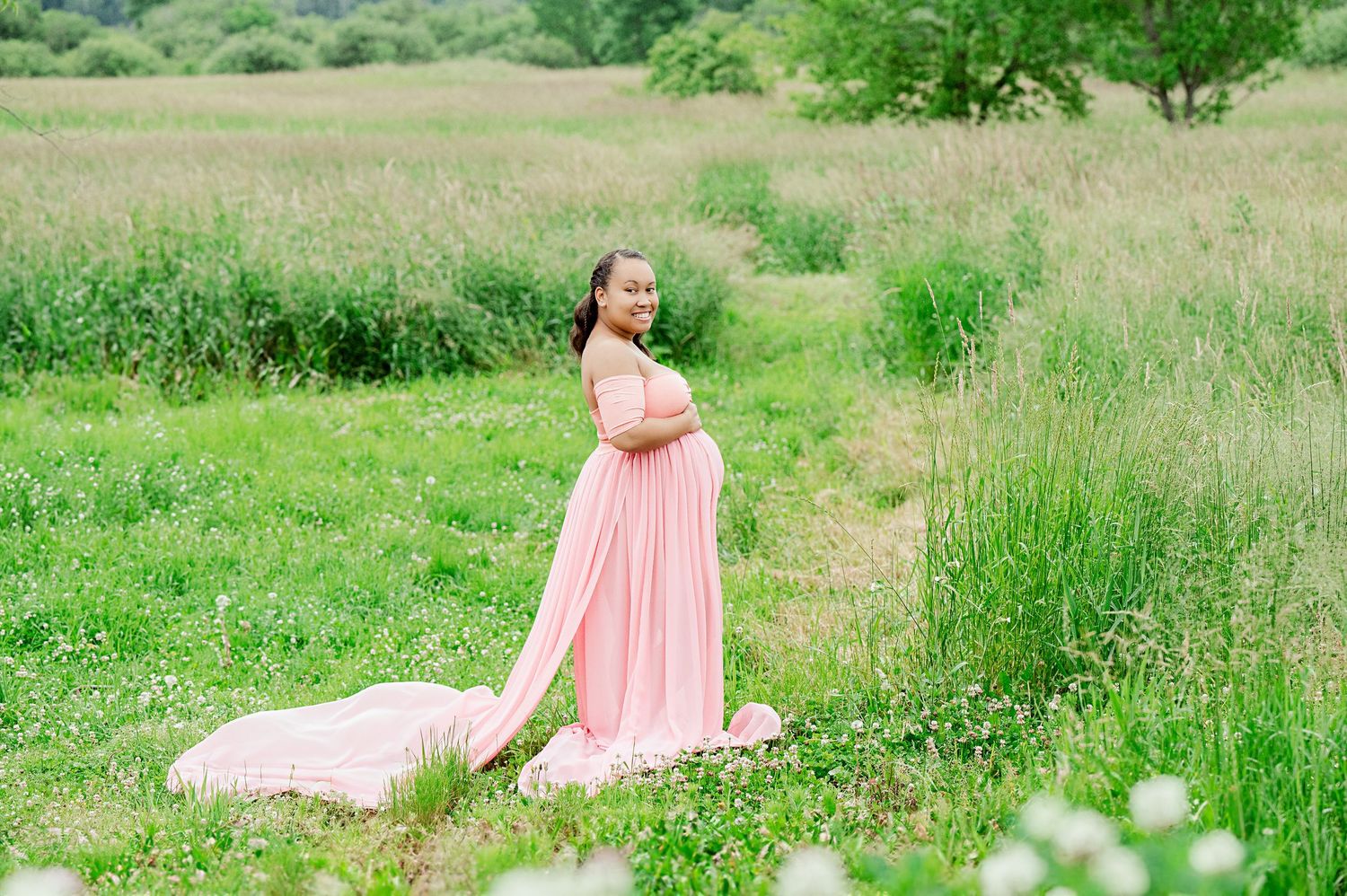 Maternity portrait of woman in flowing coral dress standing in sunlit meadow.