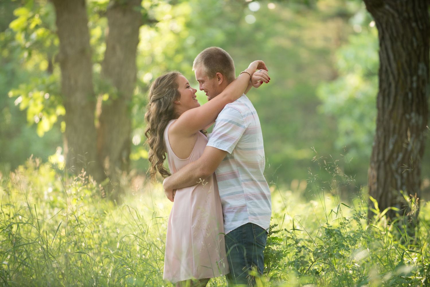 A couple embraces romantically in a sun-dappled forest setting with soft green foliage in the background.