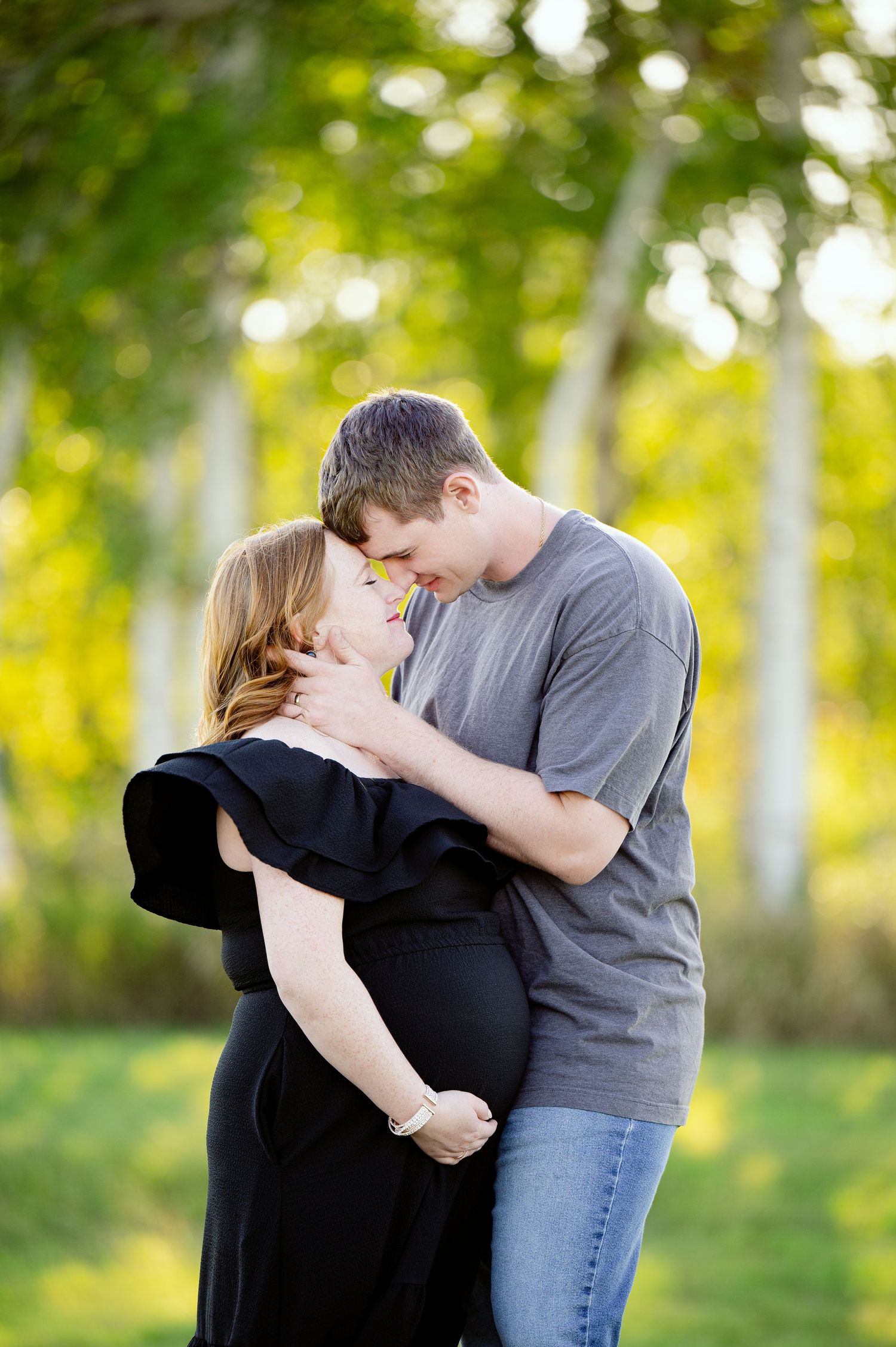 A romantic maternity portrait in a sunlit forest with couple embracing in casual attire.