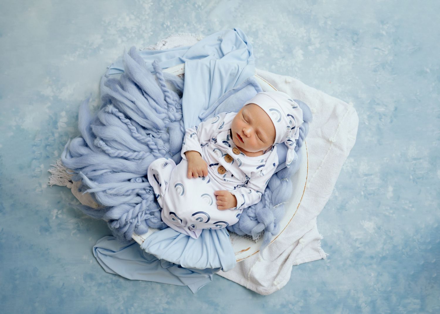 Sleeping infant in white outfit lying on flowing blue fabric against light blue backdrop.