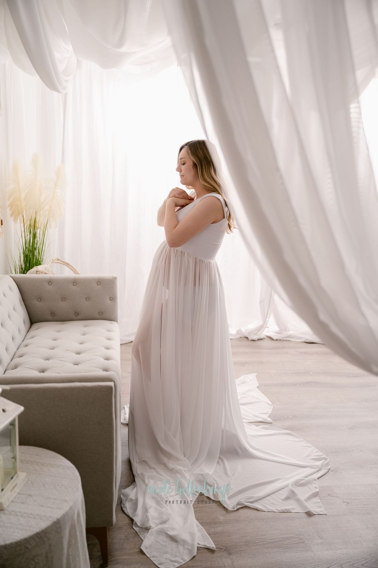 A dreamy maternity photoshoot in a bright white room with flowing curtains and a grey tufted chaise lounge.