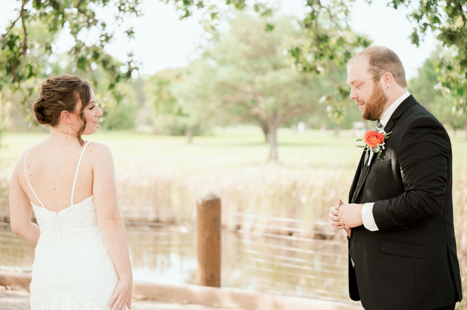 A couple celebrates their wedding outdoors by a lake with the groom in black suit and bride in white dress.