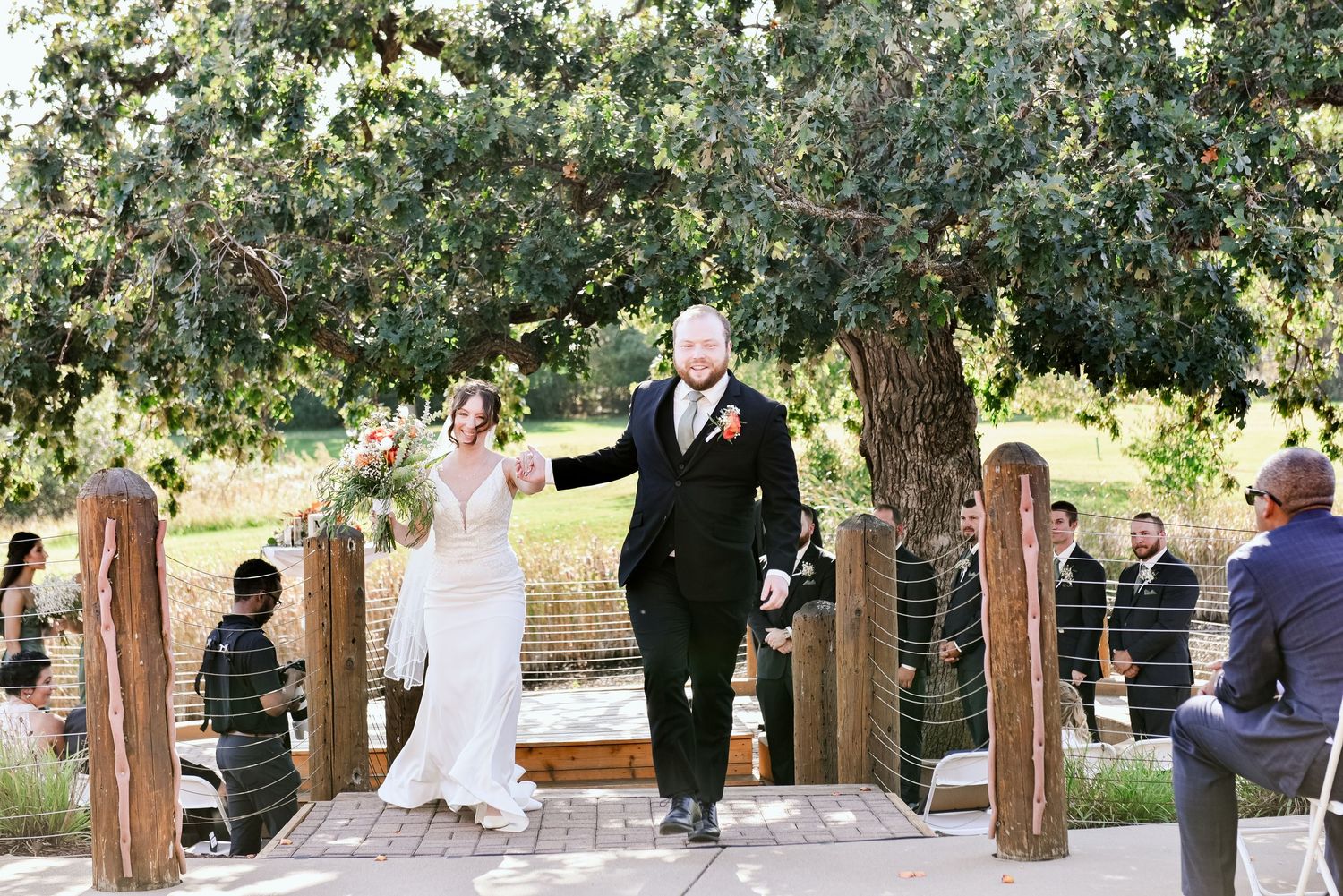Guests gather outdoors under tree-lined walkway for elegant wedding ceremony.
