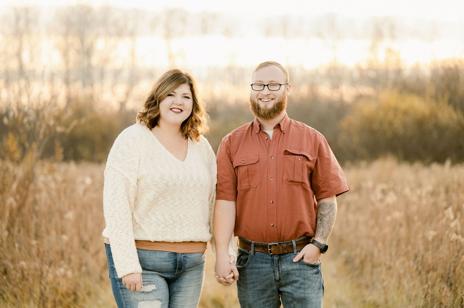 A couple stands together holding hands in a sunlit field of tall grass during golden hour.