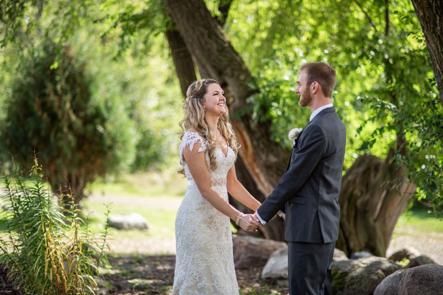 Wedding couple holds hands outdoors while sharing a joyful moment under green trees and beside natural stone landscaping.