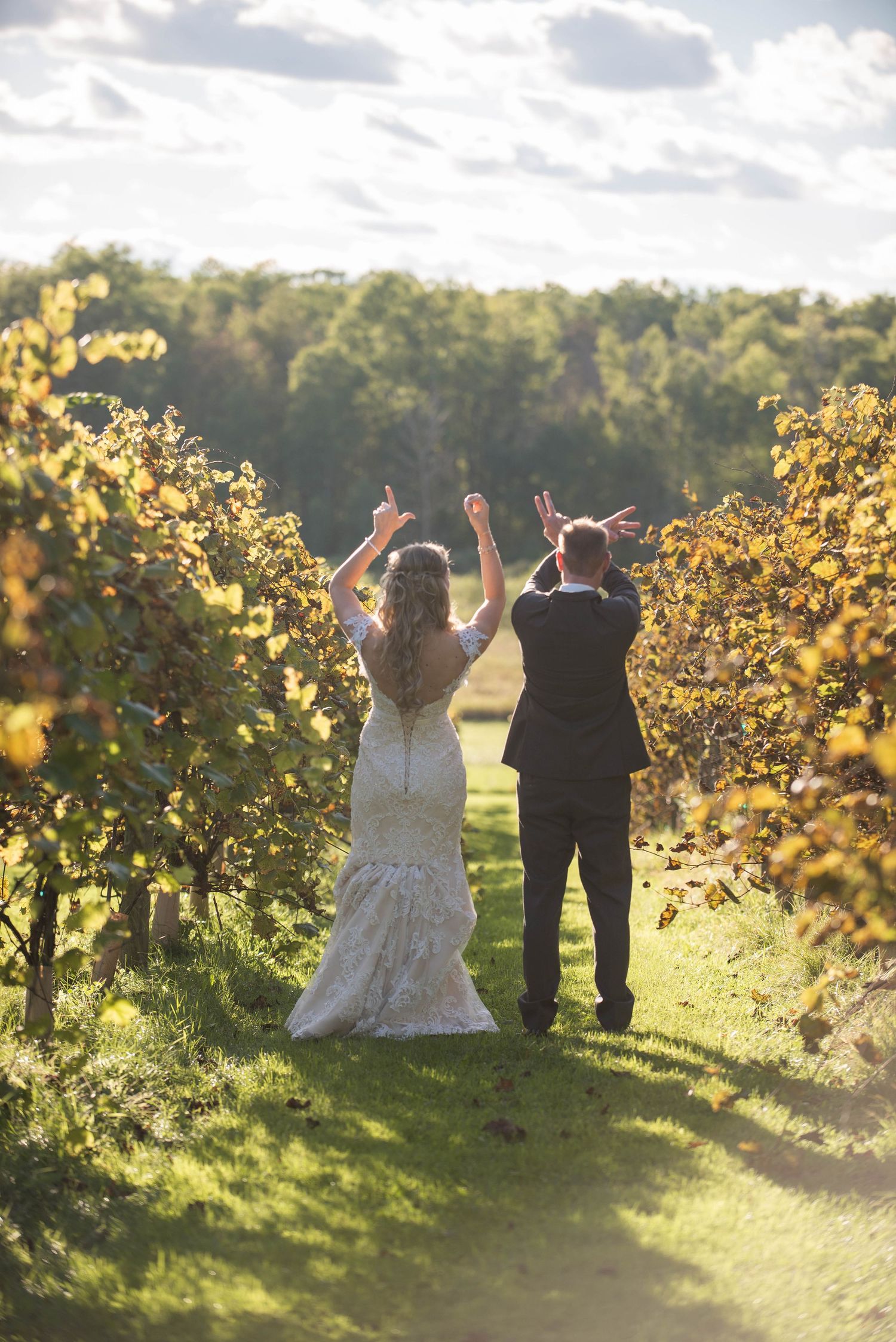 Newlyweds raise their arms in celebration among sunlit vineyard rows. Rum River Barn Milaca Minnesota Weddings