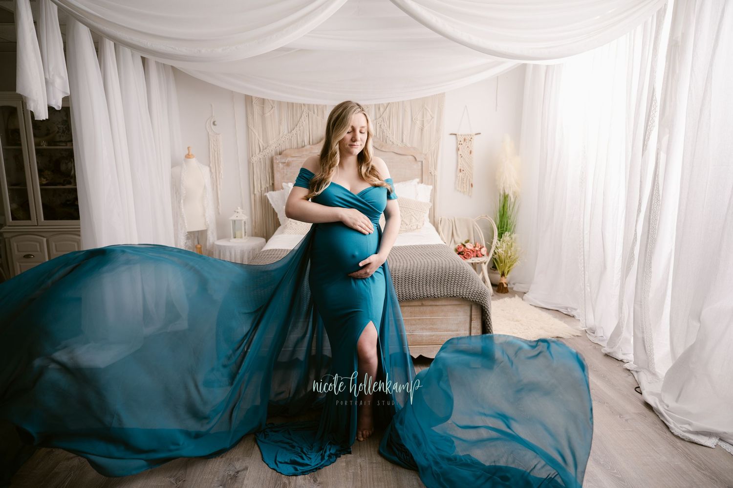 Elegant maternity photo featuring a flowing teal dress in a light and airy white draped studio setting.