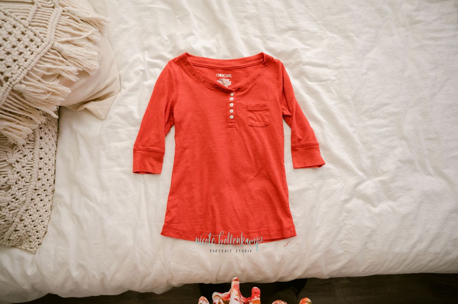 Orange cotton three-quarter sleeve henley shirt laid flat on white bedding near cream macrame decor.