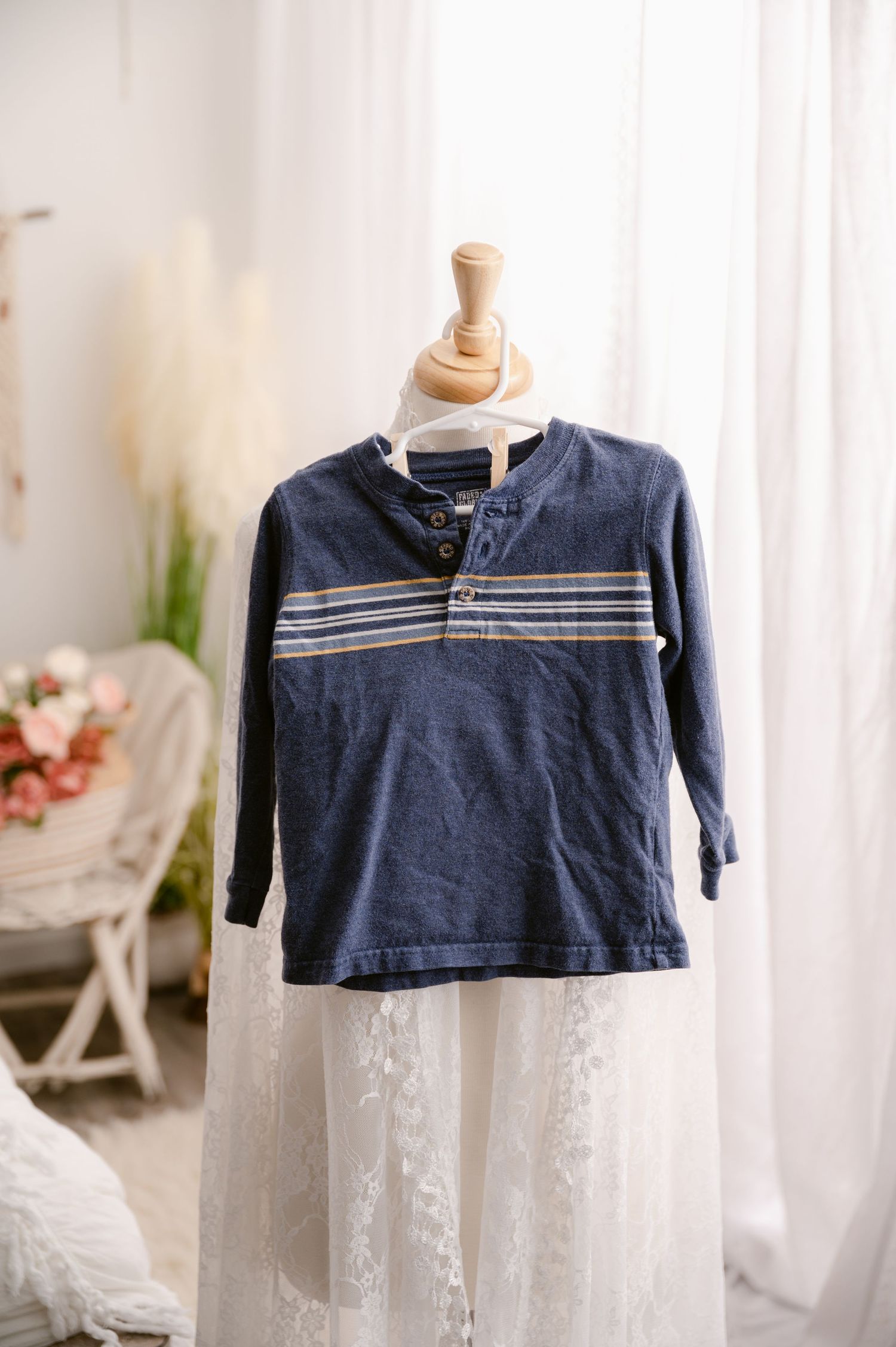 Navy blue long-sleeve henley shirt with grey stripes hangs on wooden mannequin against white curtain background.