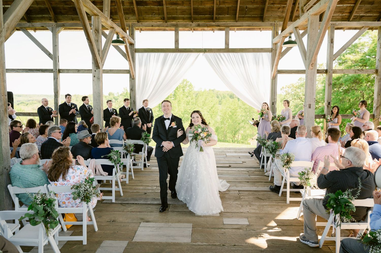 Wedding ceremony in progress inside rustic wooden pavilion with guests seated on white chairs.