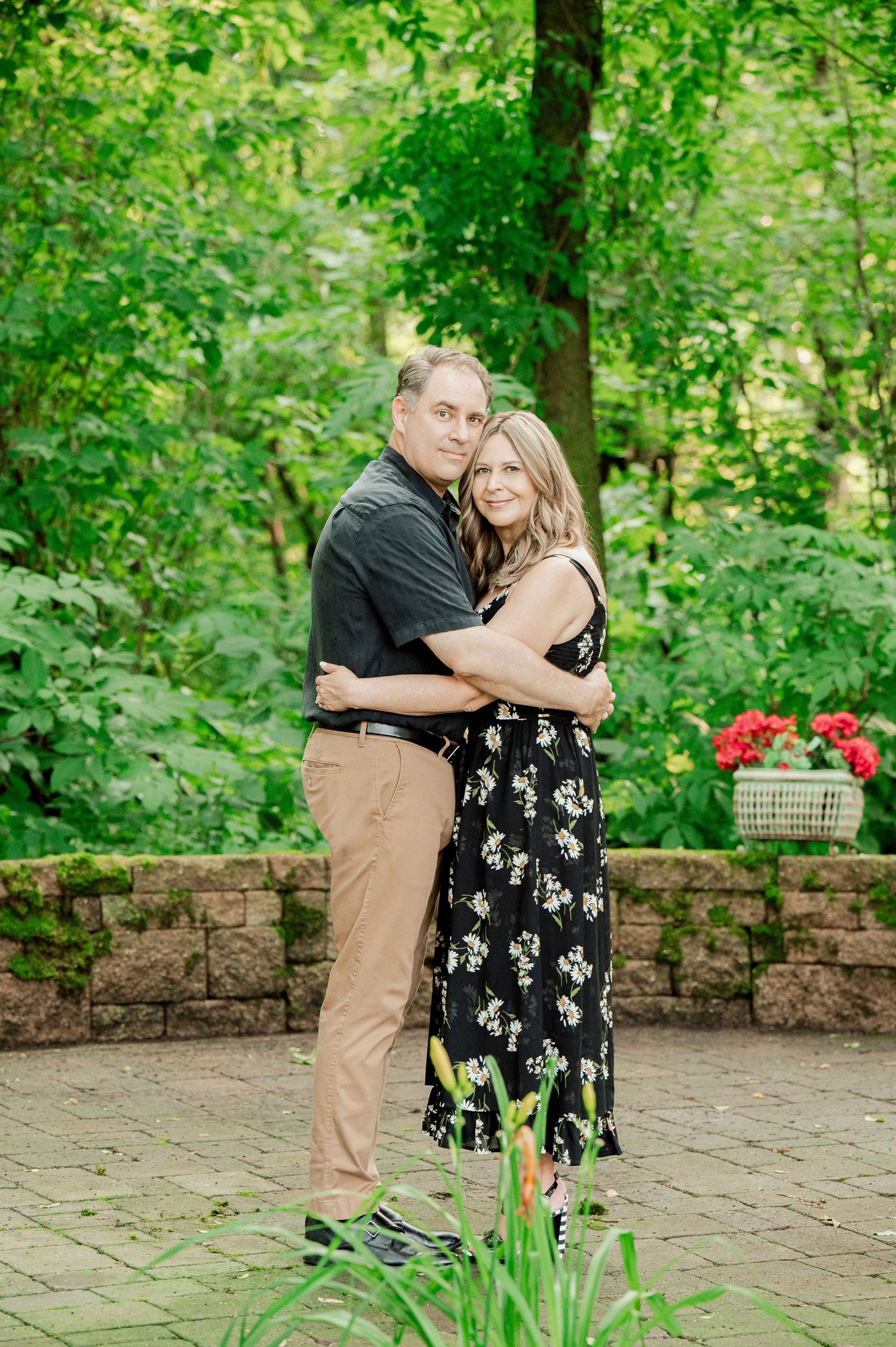 Clear Lake Family A romantic outdoor portrait of a couple embracing against a lush green forest backdrop with red flowers in a stone garden.