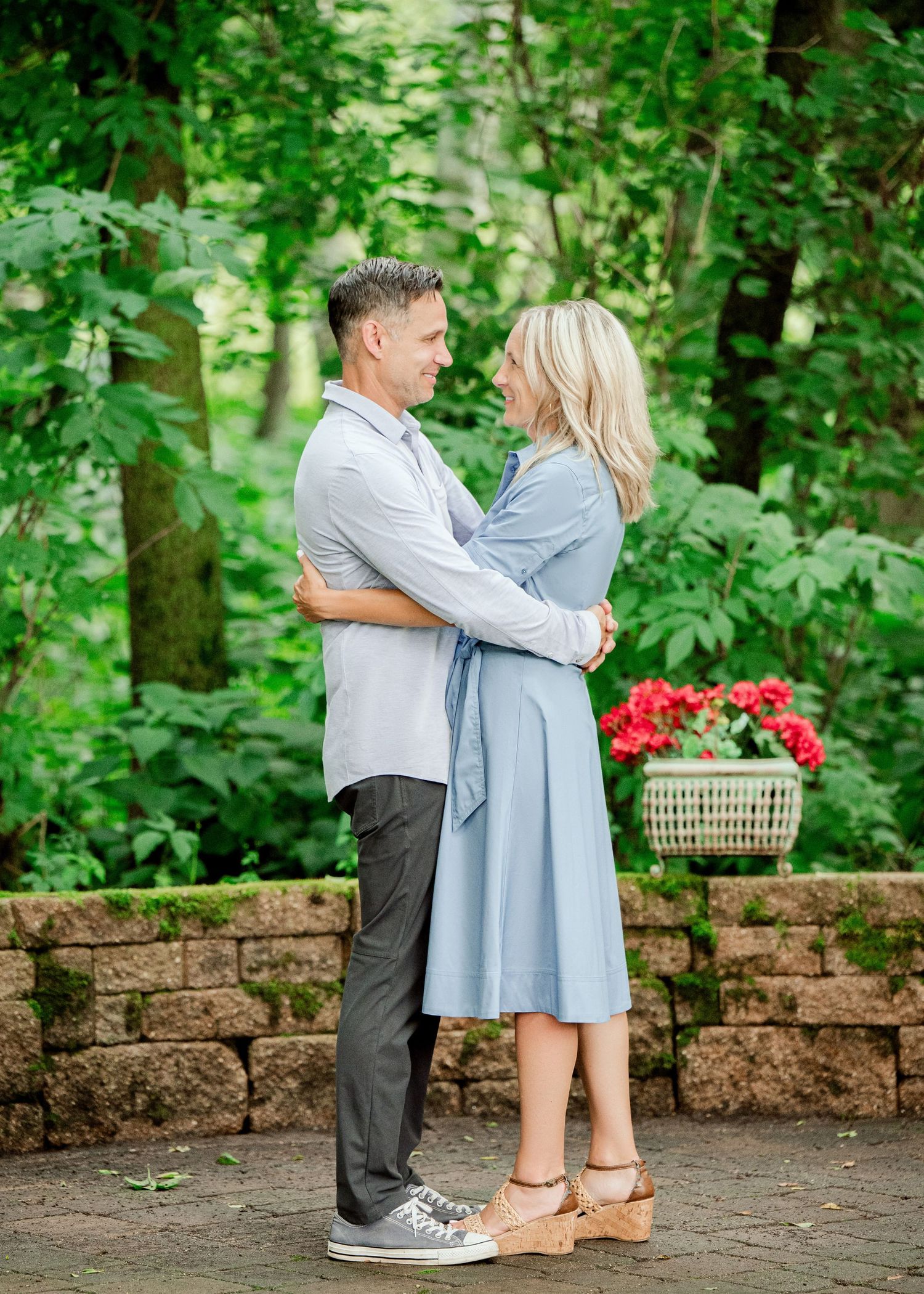Clear Lake MN Family Photos: A couple embraces on a stone patio with a basket of red flowers and lush green foliage behind them.