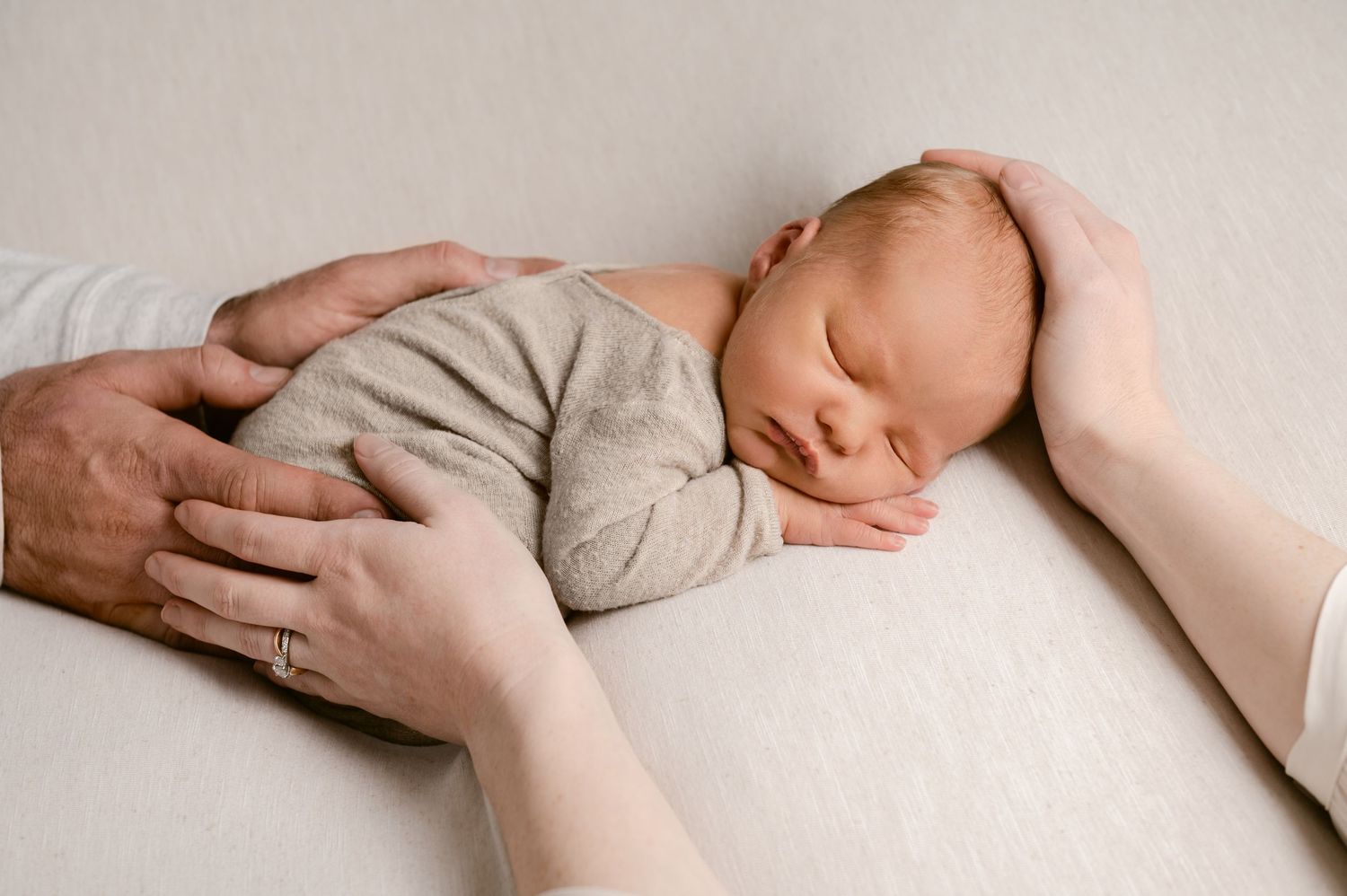 Tender moment of caring hands gently cradling sleeping newborn in soft gray onesie.