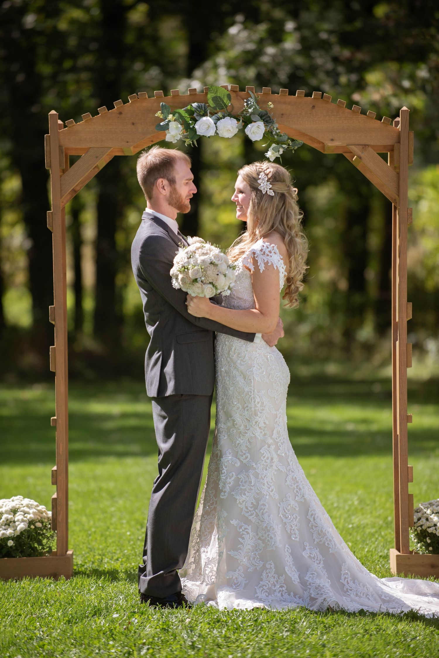 A couple shares a romantic moment under a wooden arch decorated with white flowers in a garden setting. Rum River Barn Milaca Minnesota Wedd