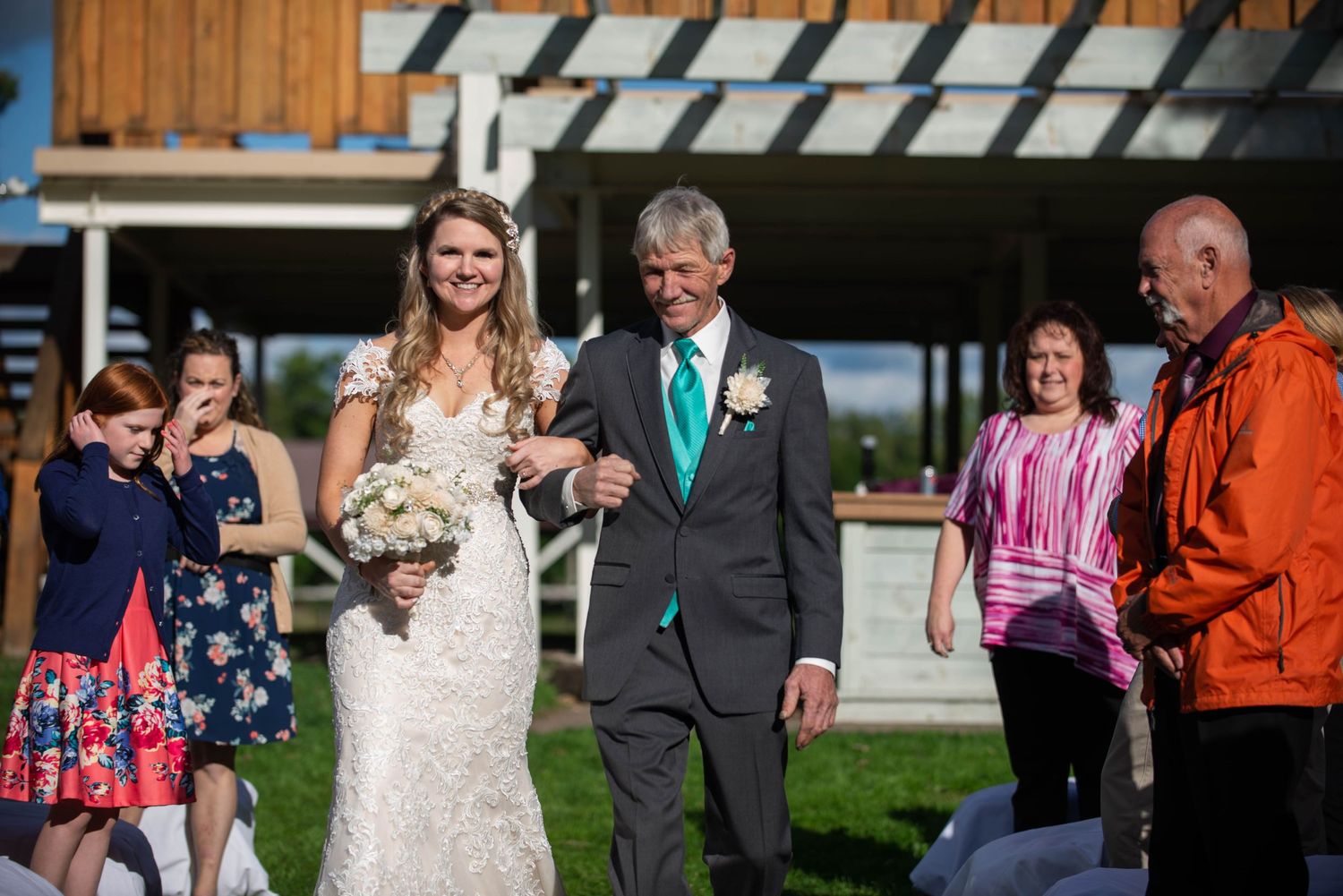 Wedding couple walking together outdoors with guests looking on near wooden building. Rum River Barn Milaca Minnesota Weddings