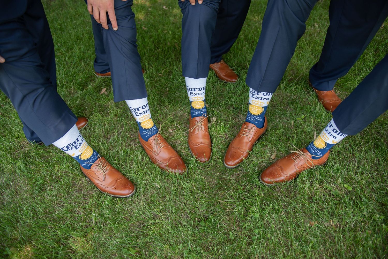 A row of groomsmen in blue suits show off their matching colorful striped socks next to leather dress shoes on green grass.
