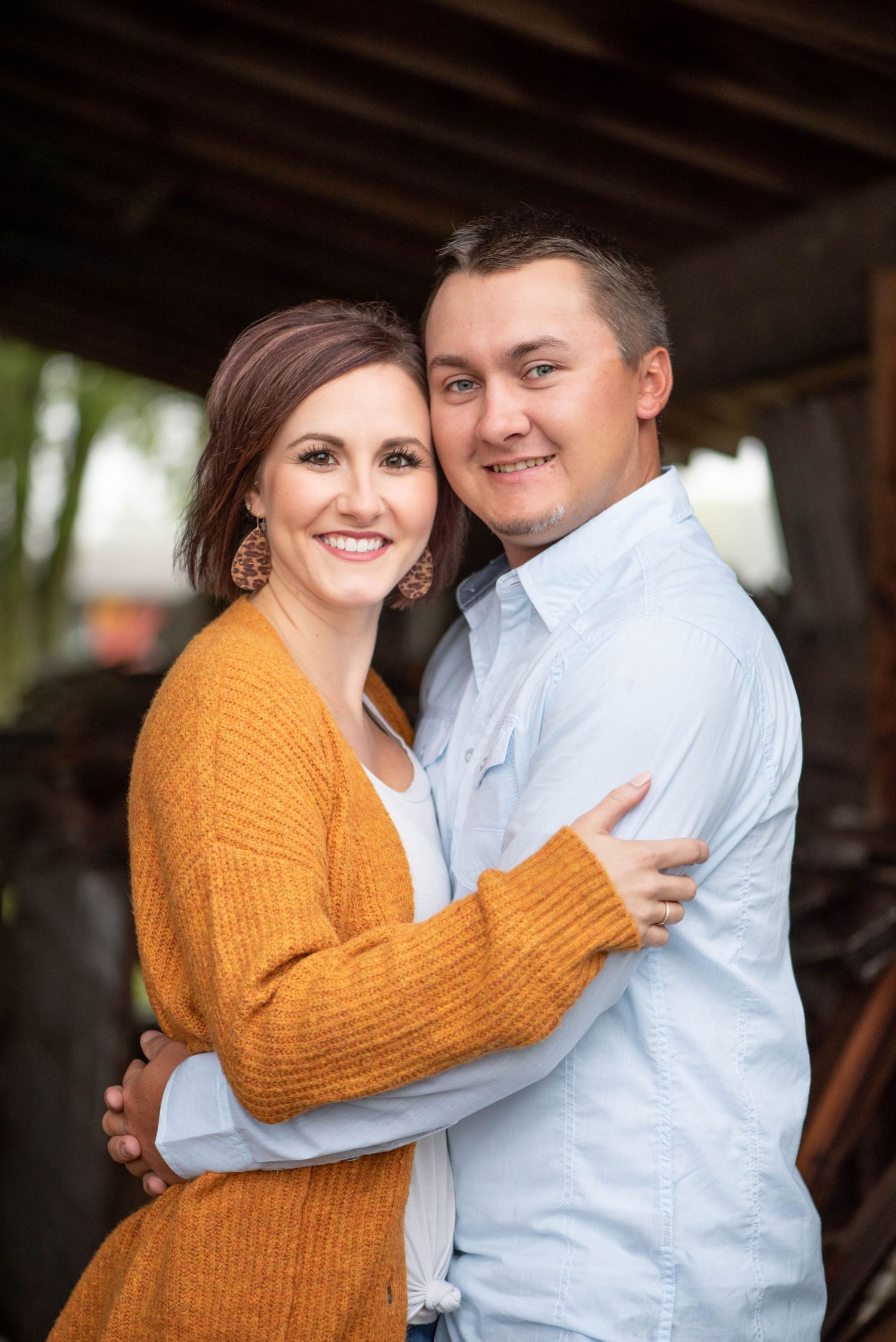 A couple embrace while smiling at the camera, with the woman wearing a mustard cardigan and the man in a light blue shirt.