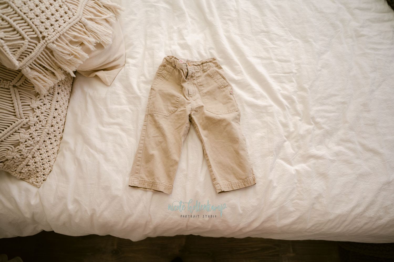 A pair of khaki pants laid flat on a white bedspread with a knitted throw blanket visible in the corner.