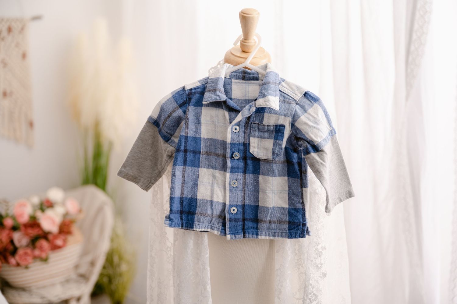 A small blue and white plaid button-down shirt with grey long sleeves hangs on a wooden hanger near flowers.