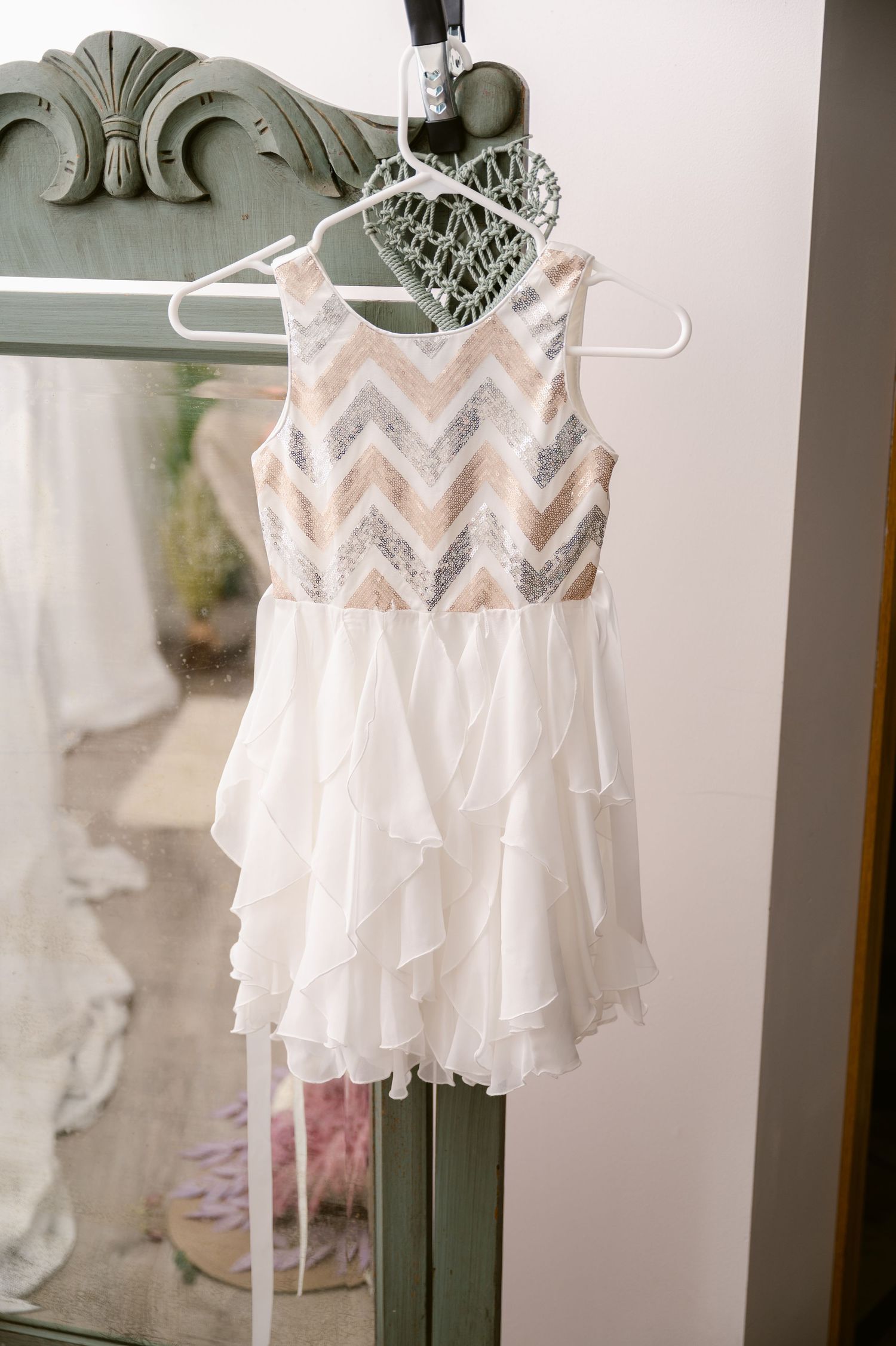 White chiffon party dress with chevron sequin bodice hangs on white hanger against gray vintage dresser.
