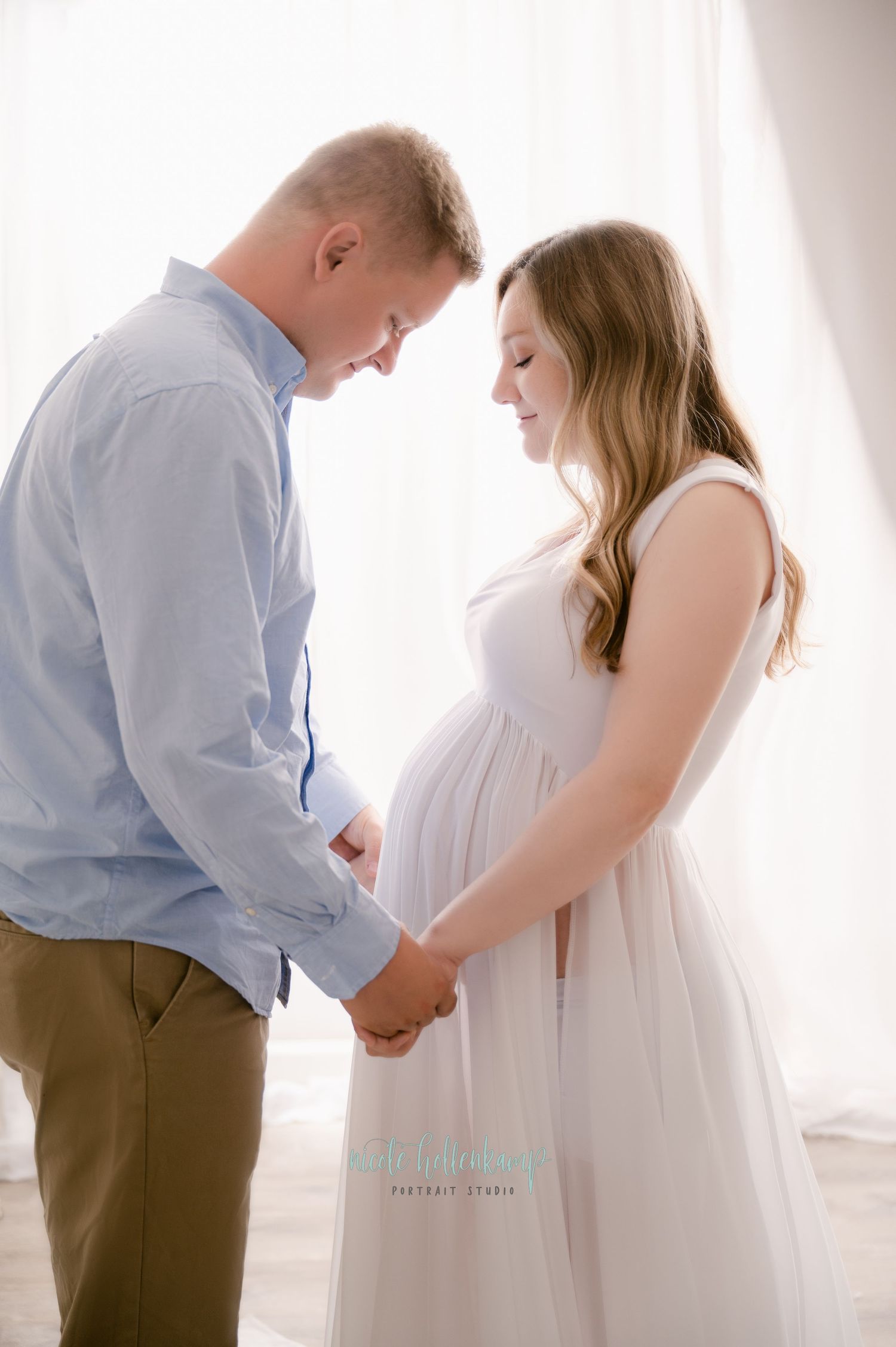 A couple stands close together in front of a sheer curtain, with the woman wearing a flowing white maternity dress.