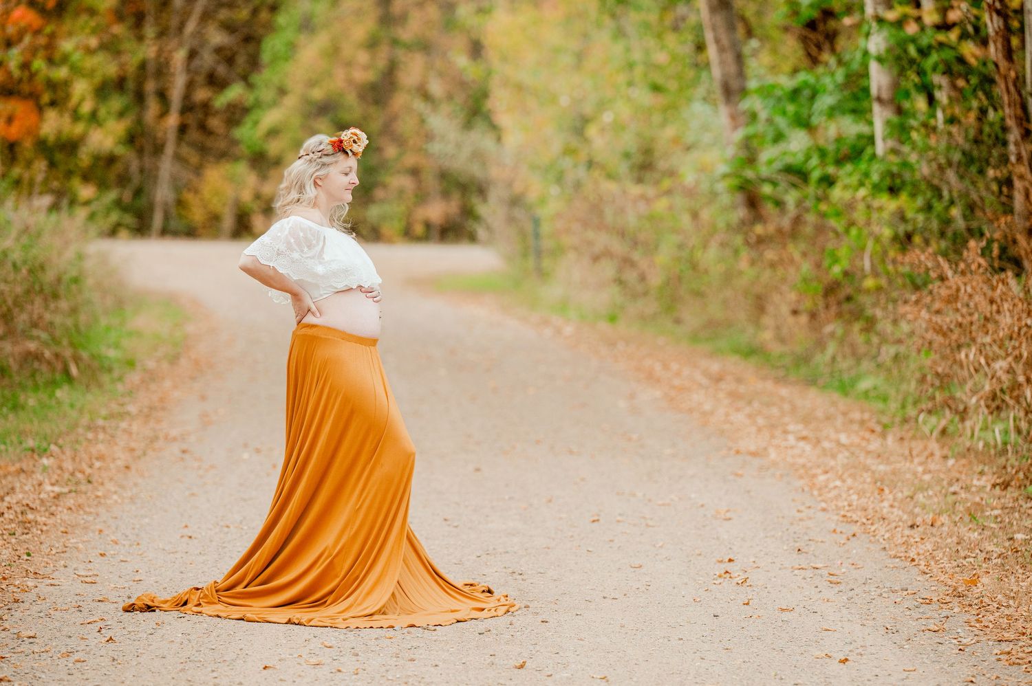 A pregnant woman in an orange flowing dress poses on a fall path during sunset.