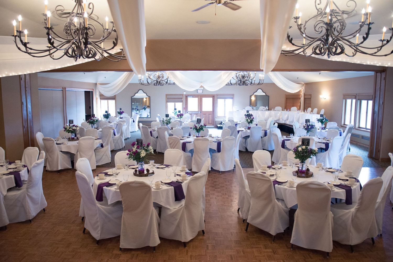 Elegant wedding reception venue with white chair covers and hanging chandeliers in a spacious ballroom. Andover wedding at Magestic Oaks Gol