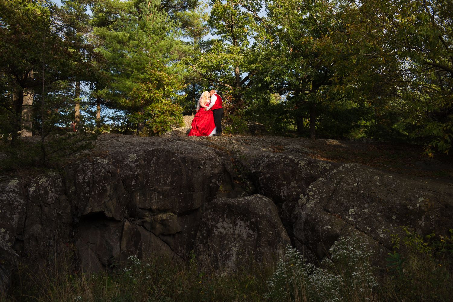 Person in red hiking gear sits atop a steep rock formation surrounded by lush green forest.