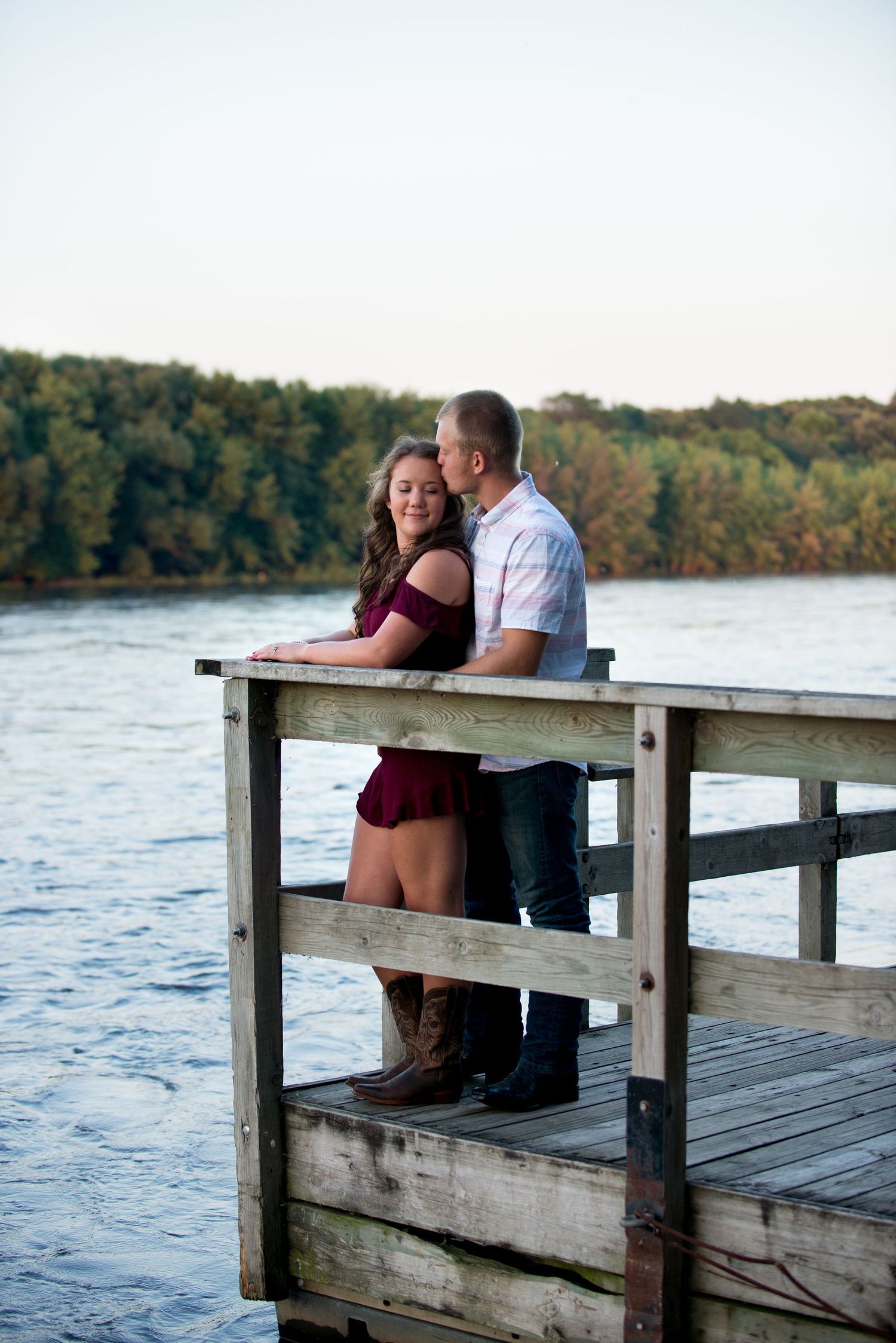 A couple sits together on a wooden dock overlooking a peaceful lake surrounded by autumn trees.