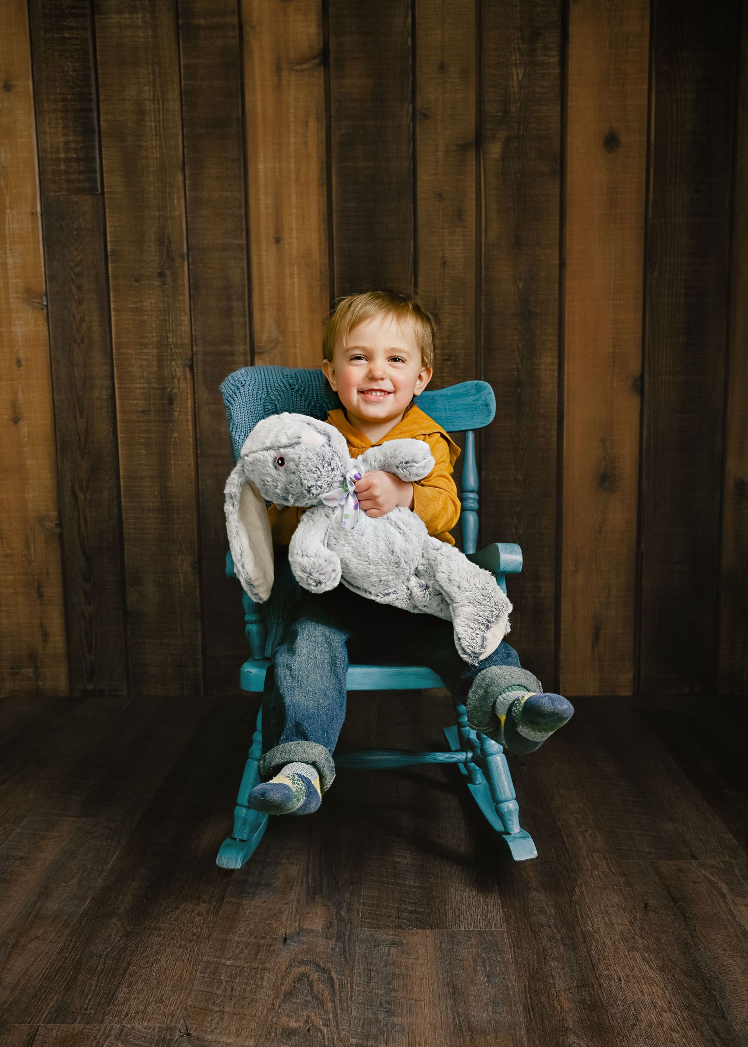 A small child sits in a turquoise rocking chair holding a plush gray bunny rabbit against a wooden wall backdrop.