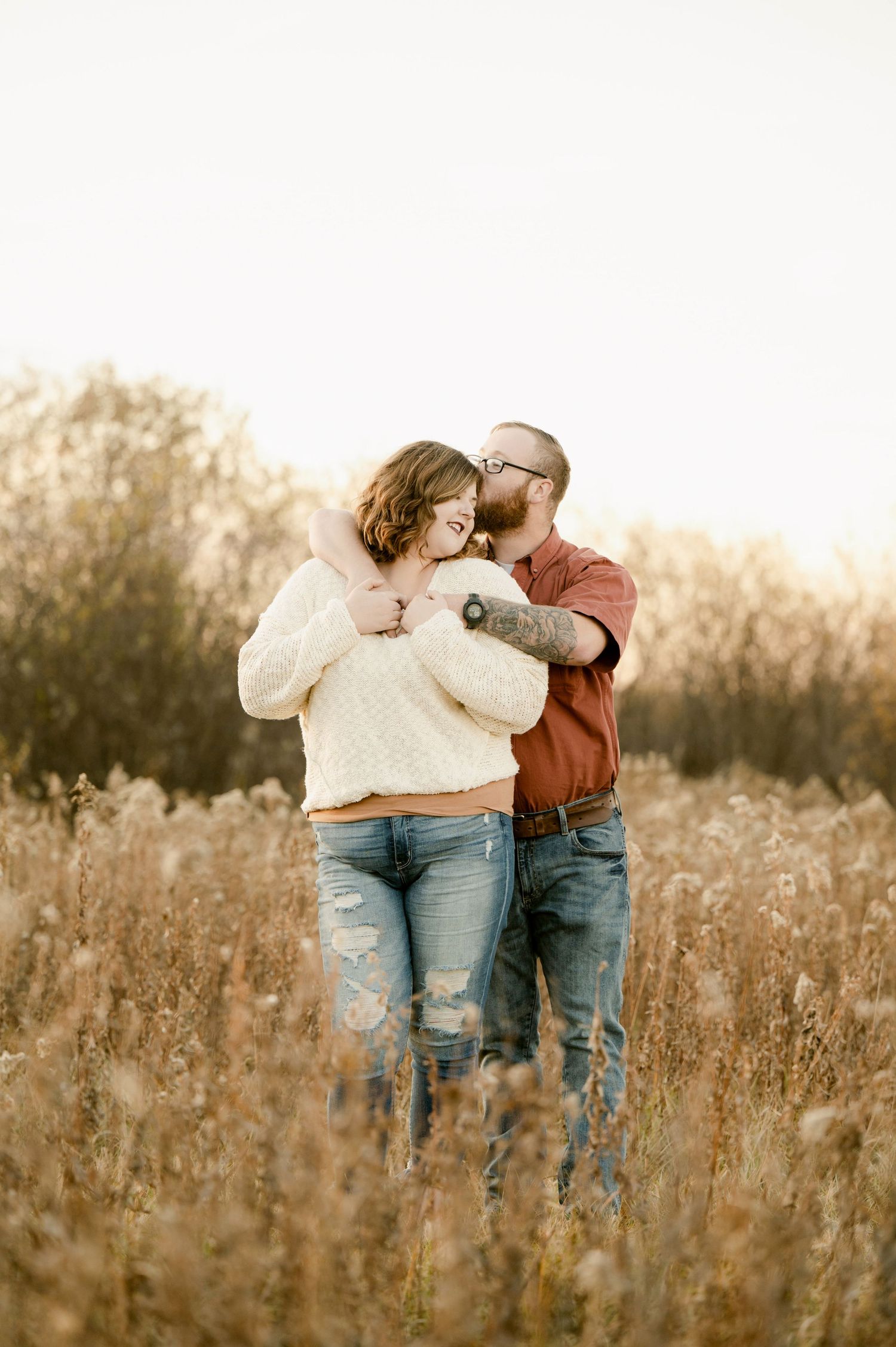 A couple embraces tenderly while standing in a golden sunlit meadow during autumn wearing casual attire. St cloud area engagement photos.