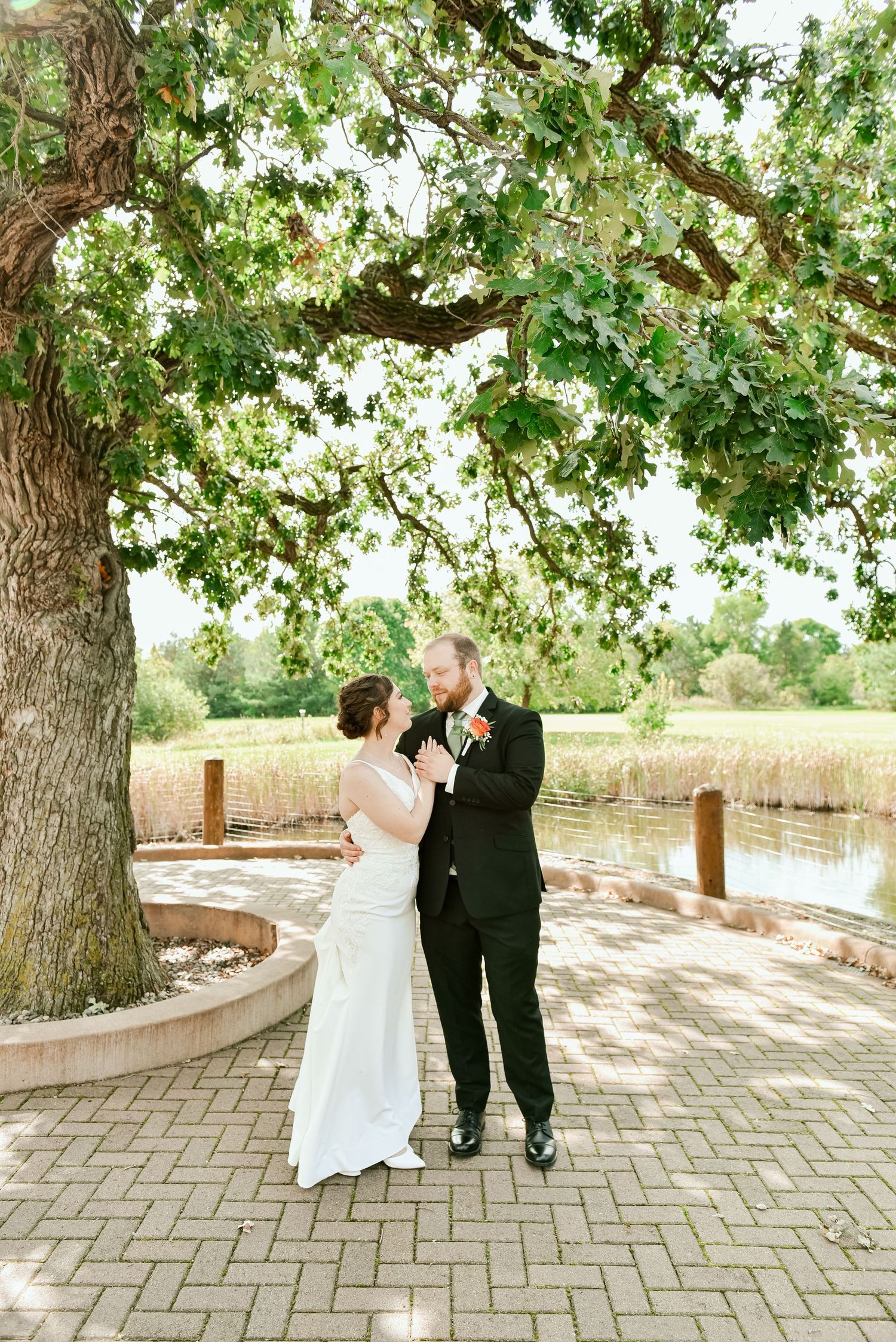 The Grands At Mulligans. A couple shares a romantic moment under a large tree beside a pond on a brick patio during their wedding ceremony.