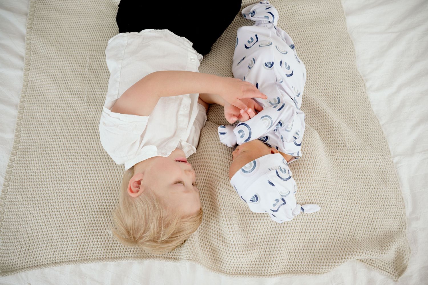 Baby in white onesie with printed pattern lying on beige knitted blanket.