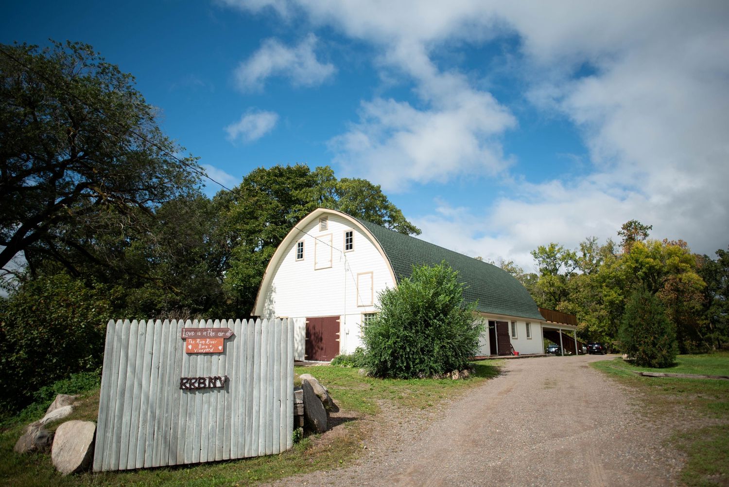 White Quonset hut barn with corrugated metal siding sits along a dirt driveway under blue skies with puffy clouds.