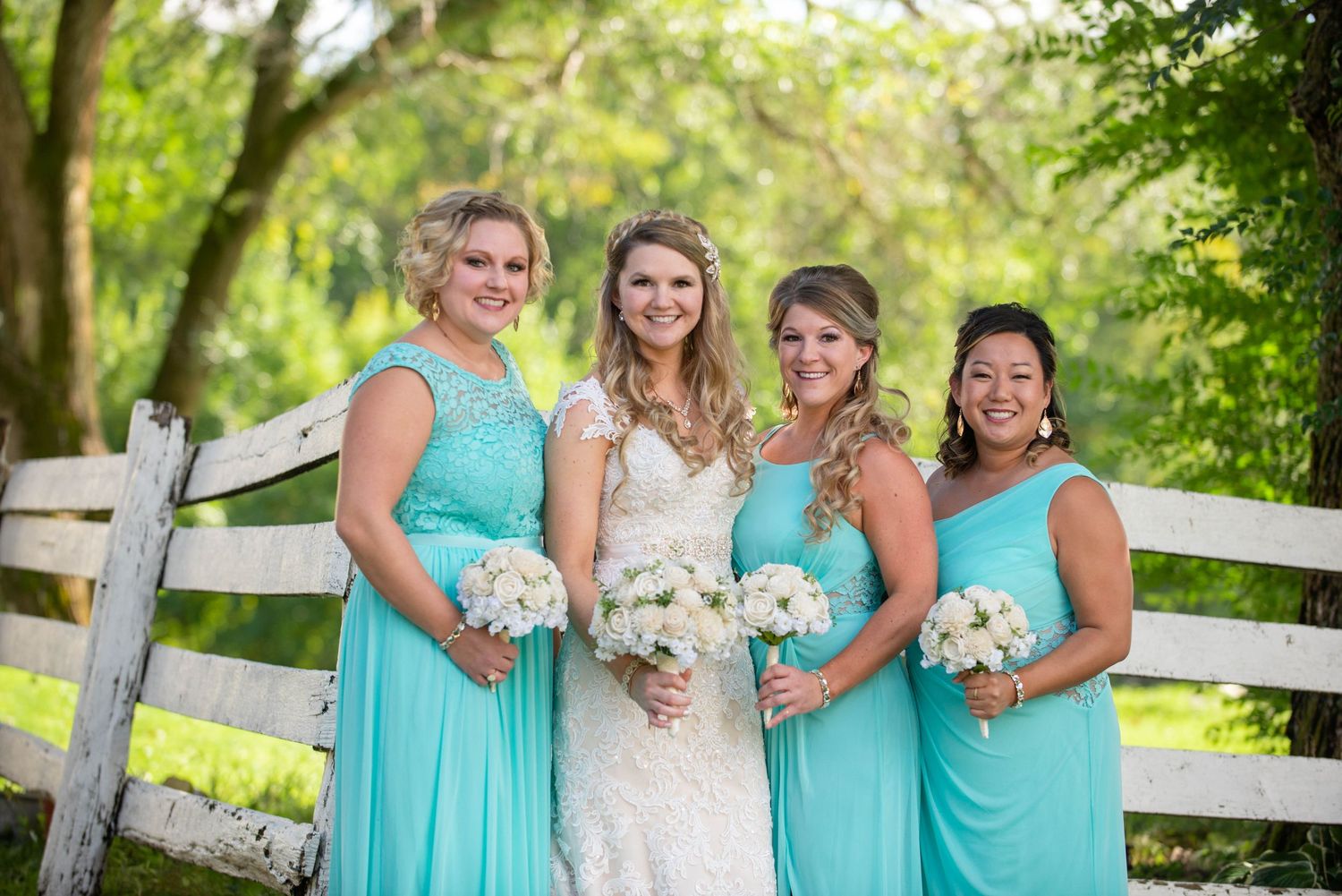 Wedding party in turquoise dresses poses by white fence in garden setting. Rum River Barn Milaca Minnesota Weddings