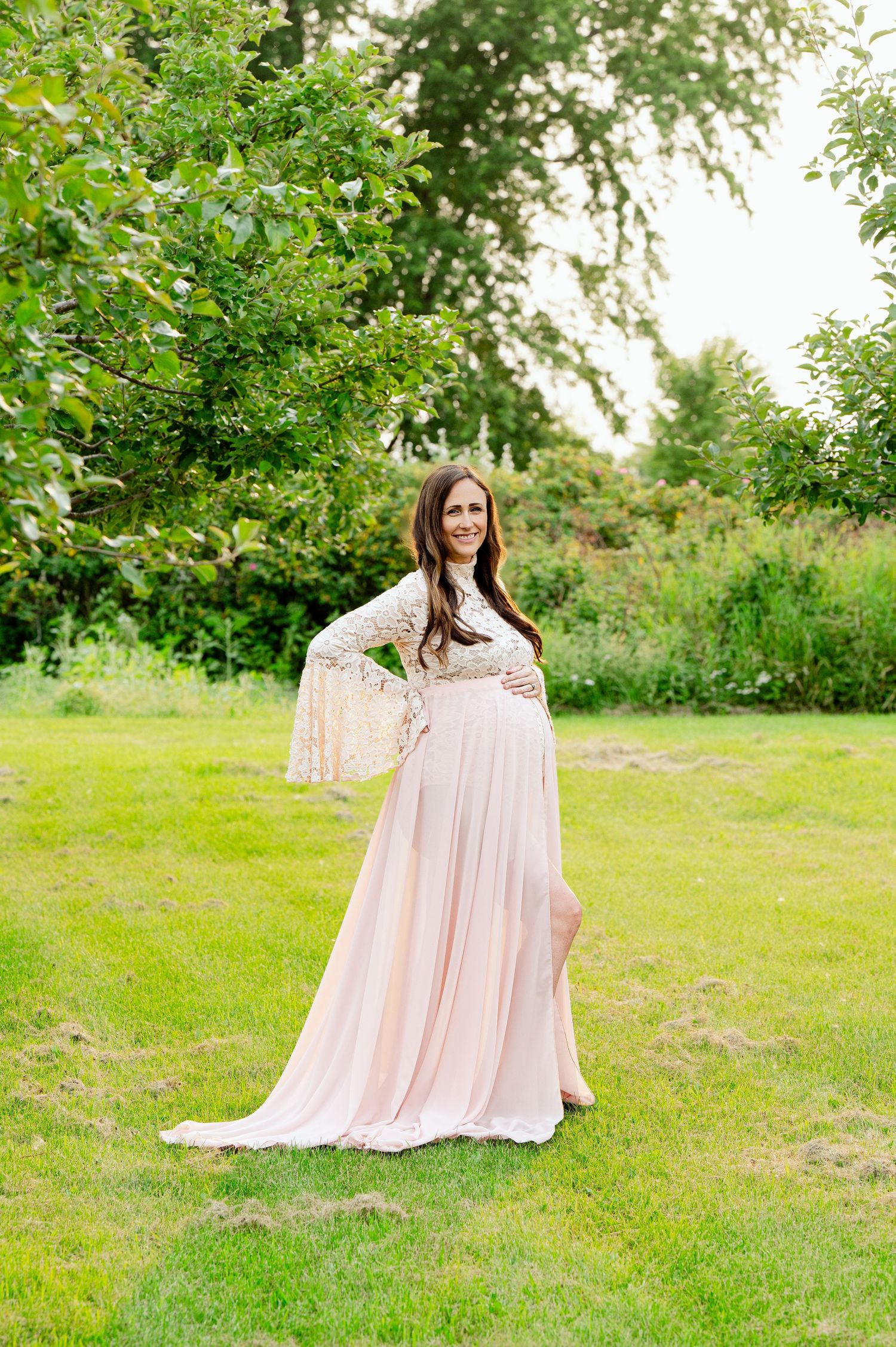 Expectant mother in flowing pink dress poses in garden setting during maternity photoshoot.