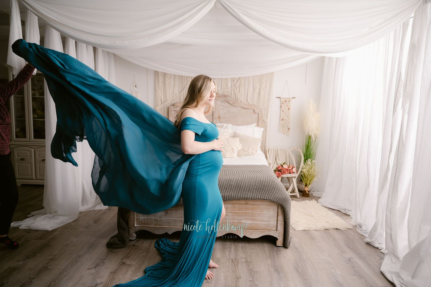 An expectant mother in a flowing teal gown stands gracefully in a draped bedroom setting with natural light.